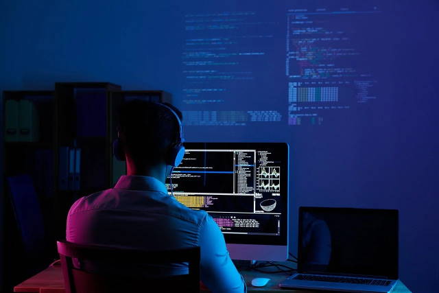 Guy with headphones working on laptop with 3 screens