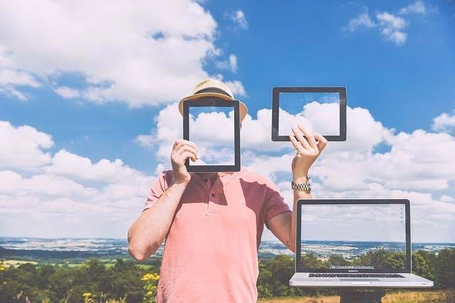 A view of the sky with a person holding up a tablet in front of their face with a cellphone and computer